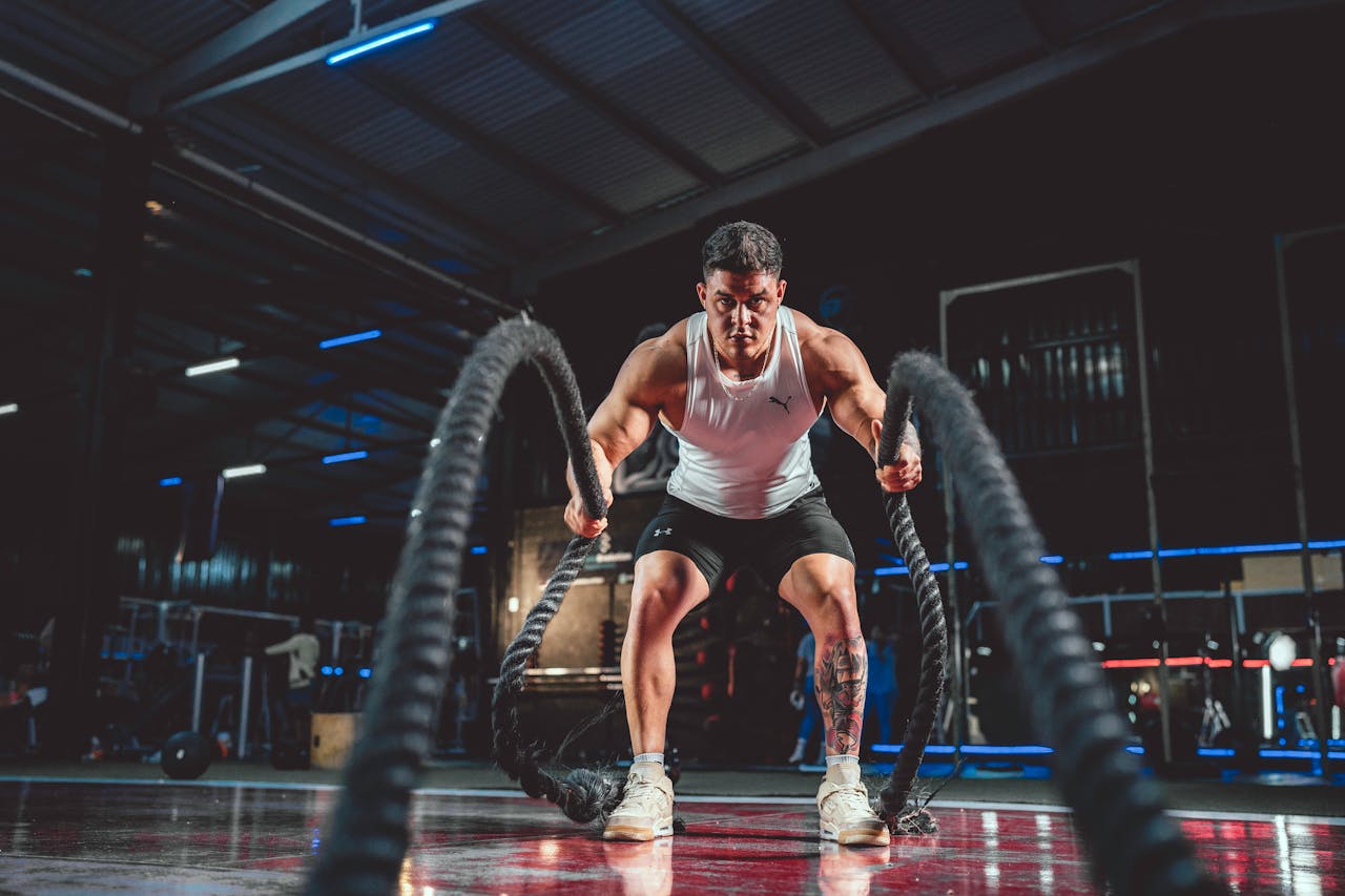 Focused man in white tank top performing battle rope exercise in a modern gym.